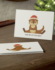 Christmas card with an otter wearing a Santa hat on a wooden surface with a Christmas tree in the background.