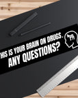 Sticker with text 'This is your brain on drugs. Any questions?' on a black surface with a ruler and pen.