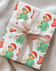 Vintage “Baby’s First Christmas” wrapping paper featuring a smiling baby in a mint green romper and Santa hat holding a white bunny plush, surrounded by red Christmas stockings and holly, retro storybook style.