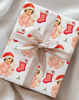 Vintage “Baby’s First Christmas” wrapping paper featuring a smiling brunette baby girl in a pink romper and Santa hat holding a white bunny plush and candy cane, surrounded by red Christmas stockings and holly, retro storybook illustration style.