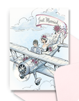 Vintage wedding greeting card with bride and groom flying in a biplane with Just Married banner trailing behind. A pink envelope is laying in the background.