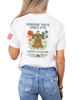 Woman wearing a white t-shirt with a graphic design of a liberty bell and flowers including text that says ringing true since 1776 with an american flag sleeve back view