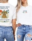 Two views of a white t-shirt with 'Ringing True Since 1776' design, featuring a bell and floral elements.