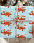 christmas wrapping paper featuring santa flying a biplane through blue sky.