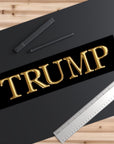 Gold 'TRUMP' text on a black background with a ruler and pens on a wooden surface