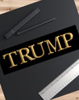 Gold 'TRUMP' text on a black background with pens and a ruler on a wooden surface