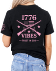 Black t-shirt with pink graphic that includes crossed muskets american flag and stars with text on the back, that says 1776 vibes trust in god