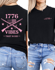 Black t-shirt with pink graphic that includes crossed muskets american flag and stars with text on the back, that says 1776 vibes trust in god displayed from front and back views.