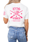 Woman wearing a white t-shirt with pink text that says 1776 vibes trust in God with muskets and an american flag on the back