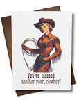 A retro style greeting card with a Western theme, featuring an illustration of a woman in a cowboy hat with a lasso. The text on the card reads 'You've lassoed another year, cowboy!'.