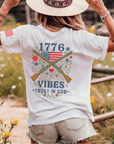 woman wearing a white t-shirt with patriotic design crossed muskets floral adorned with a betsy ross flag trust in god text