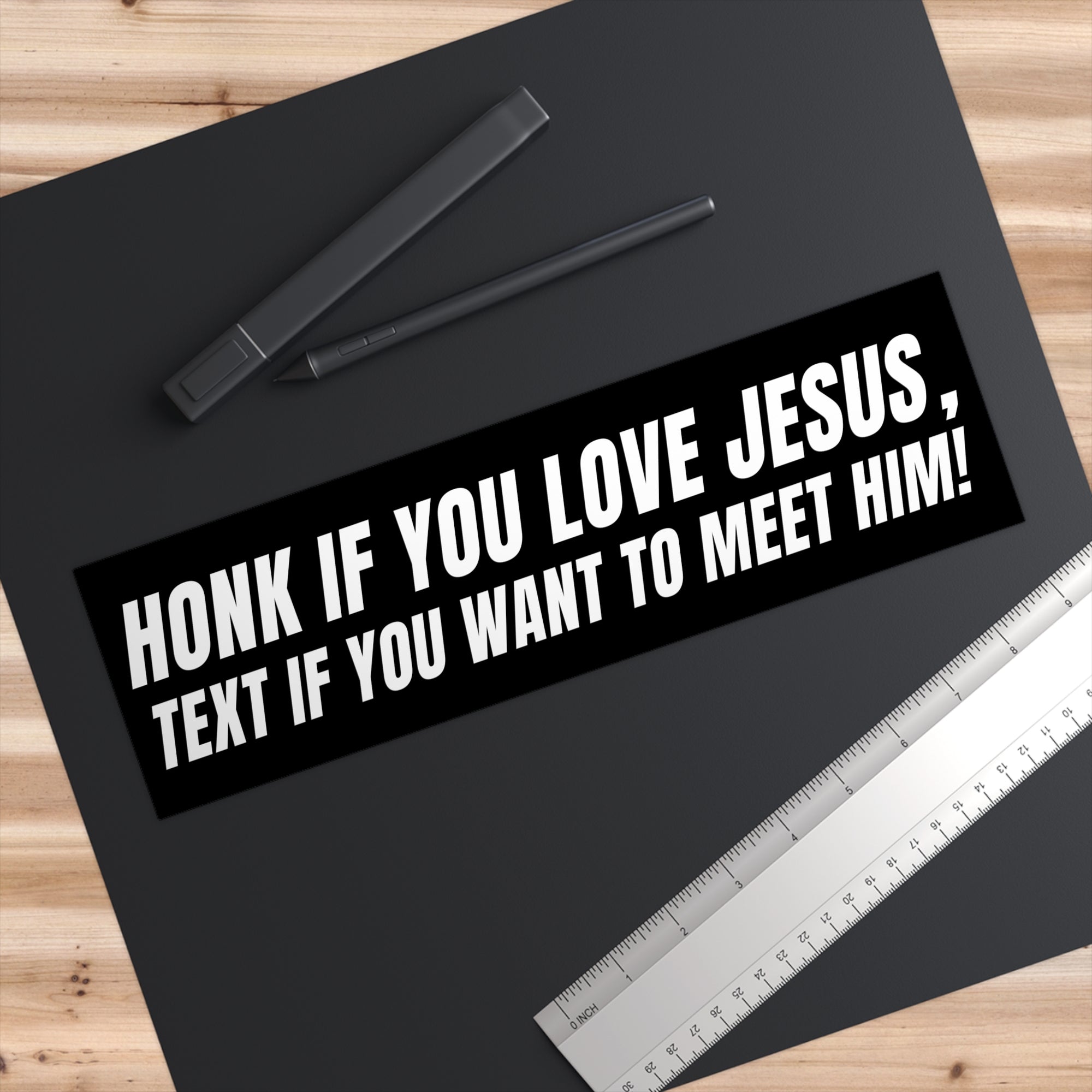 Black bumper sticker with white text on a black surface with pens and a ruler.