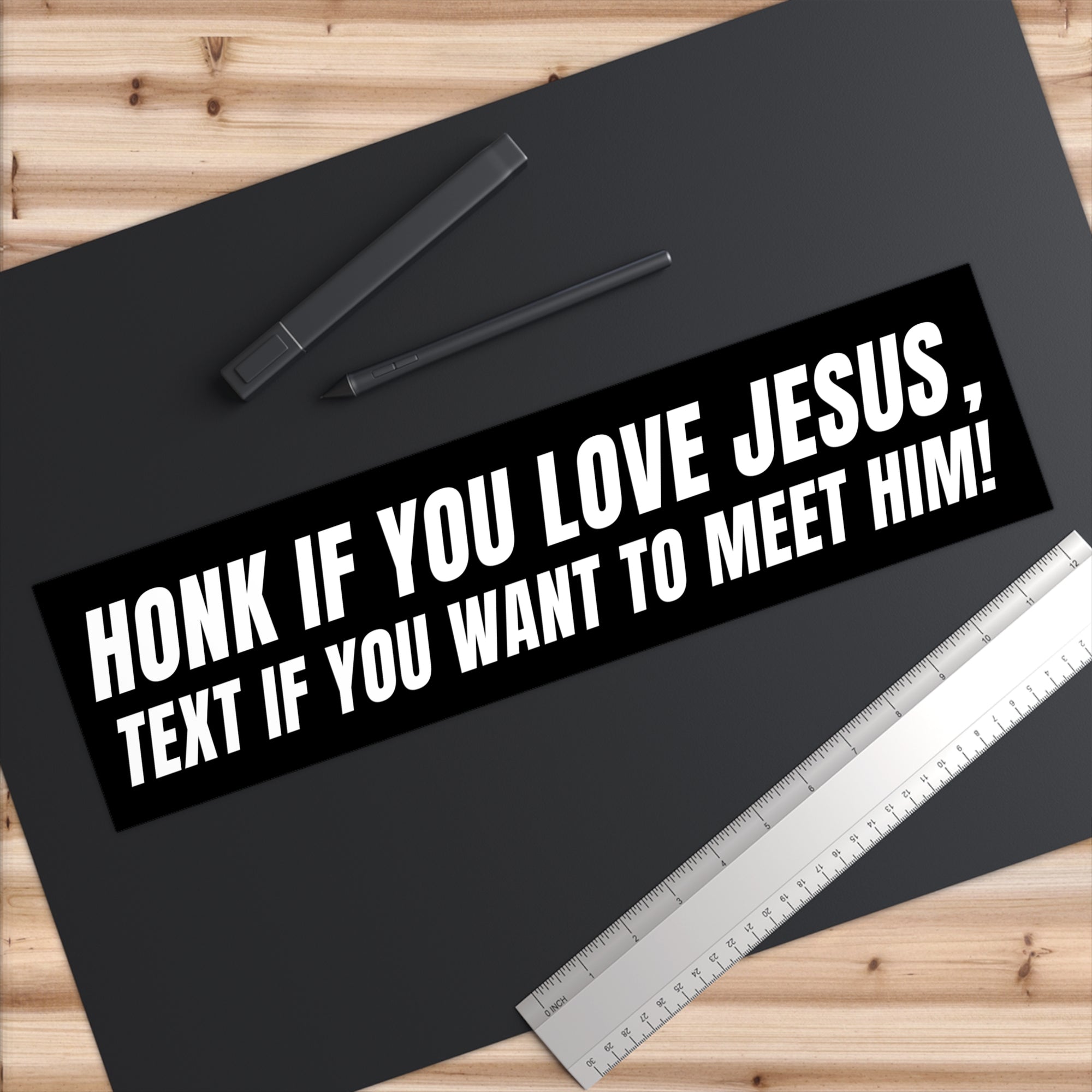 Black bumper sticker with white text on a black surface with a pen and ruler.