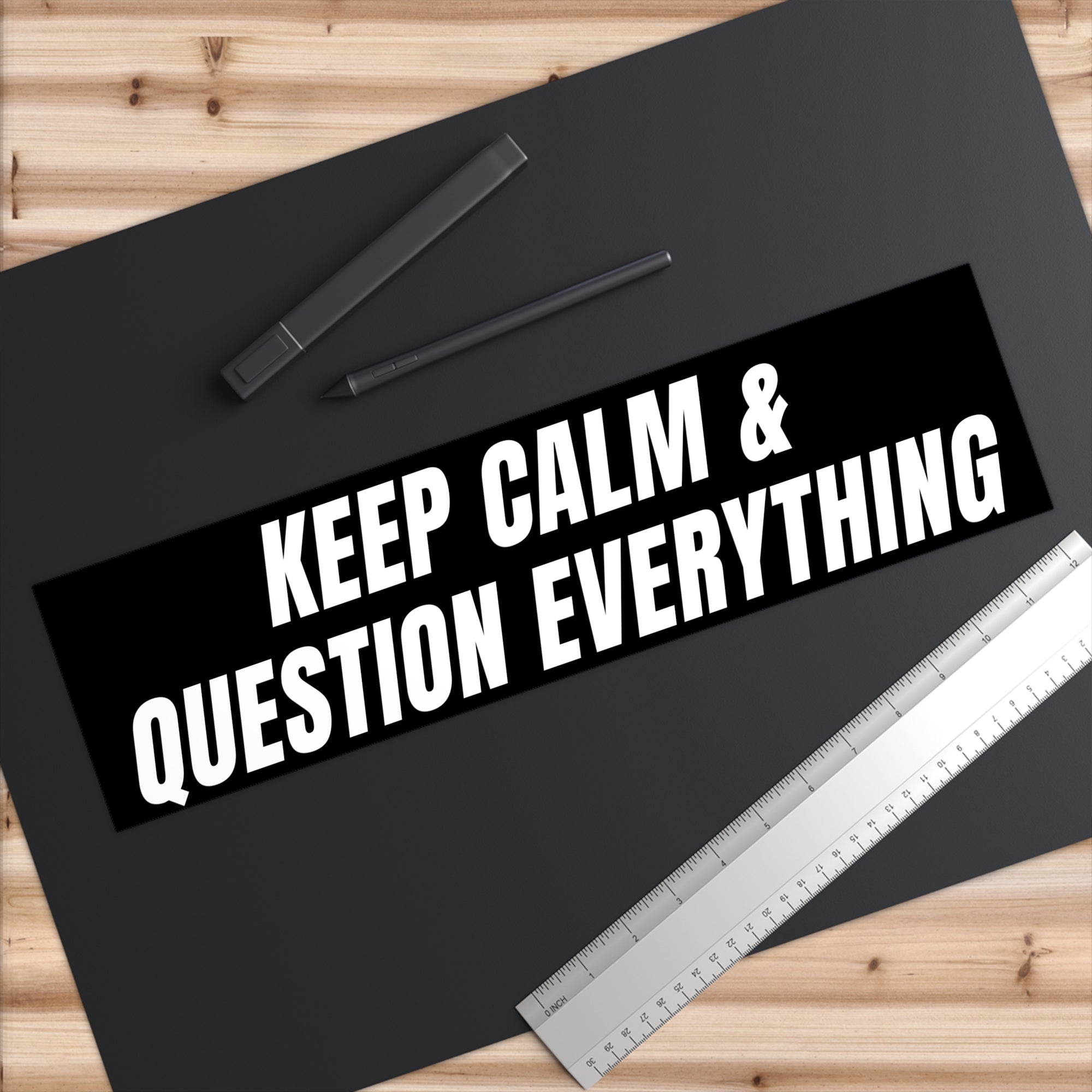 Black mat with 'KEEP CALM & QUESTION EVERYTHING' text, pen, and ruler on a wooden surface