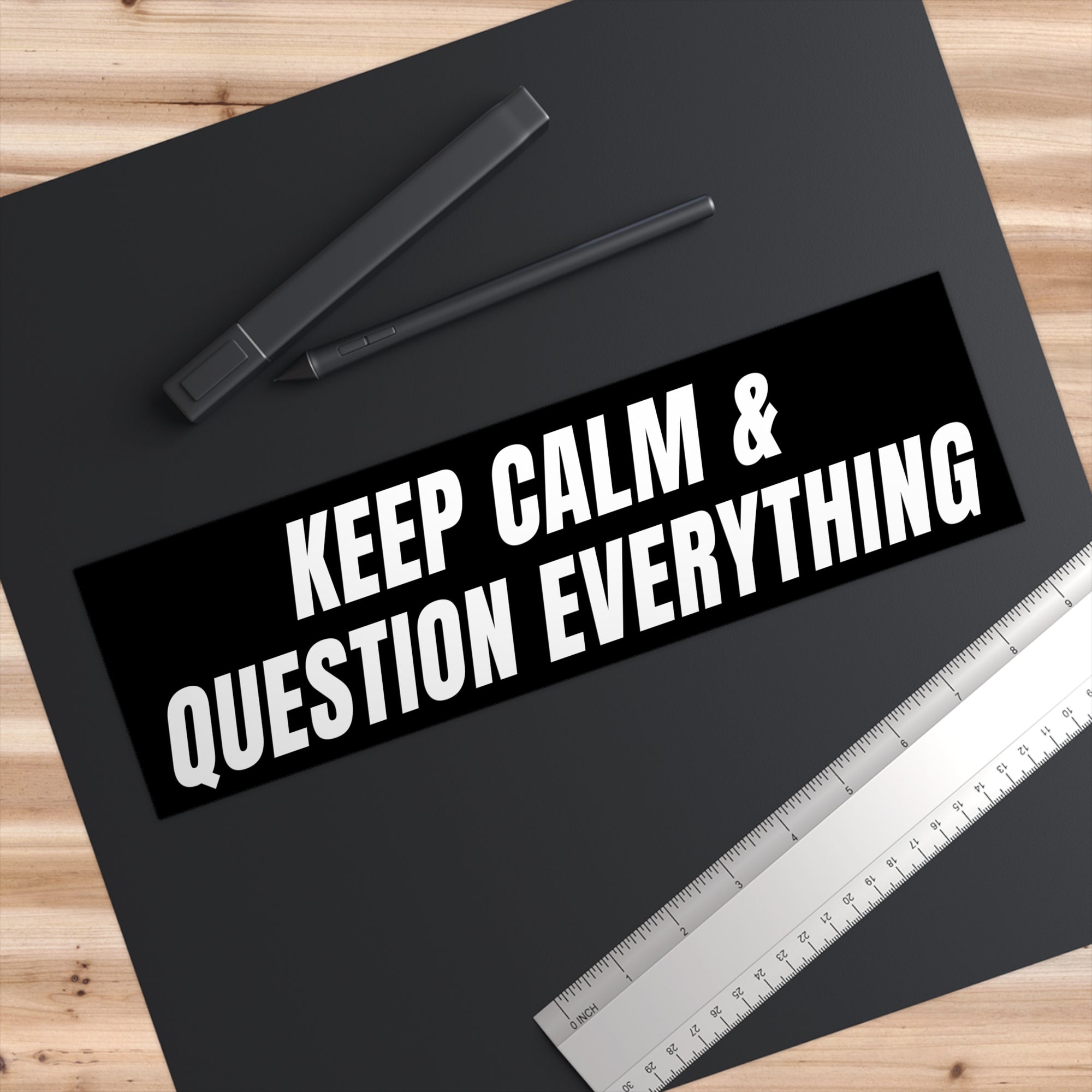 Black mat with 'Keep Calm & Question Everything' text, pen, and ruler on a wooden surface