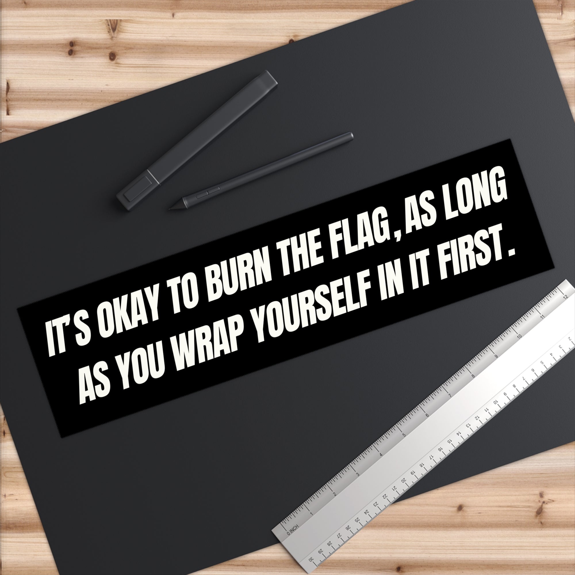 Black bumper sticker with white text on a black surface with a ruler and pen.