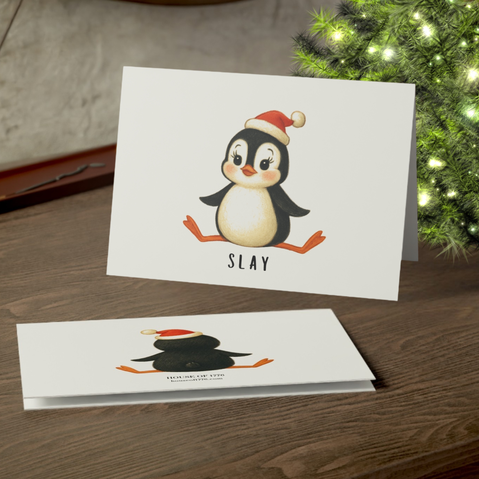Christmas card with a penguin wearing a Santa hat and the word 'SLAY' on a wooden surface with a Christmas tree in the background.