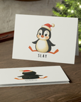 Christmas card with a penguin wearing a Santa hat and the word 'SLAY' on a wooden surface with a Christmas tree in the background.