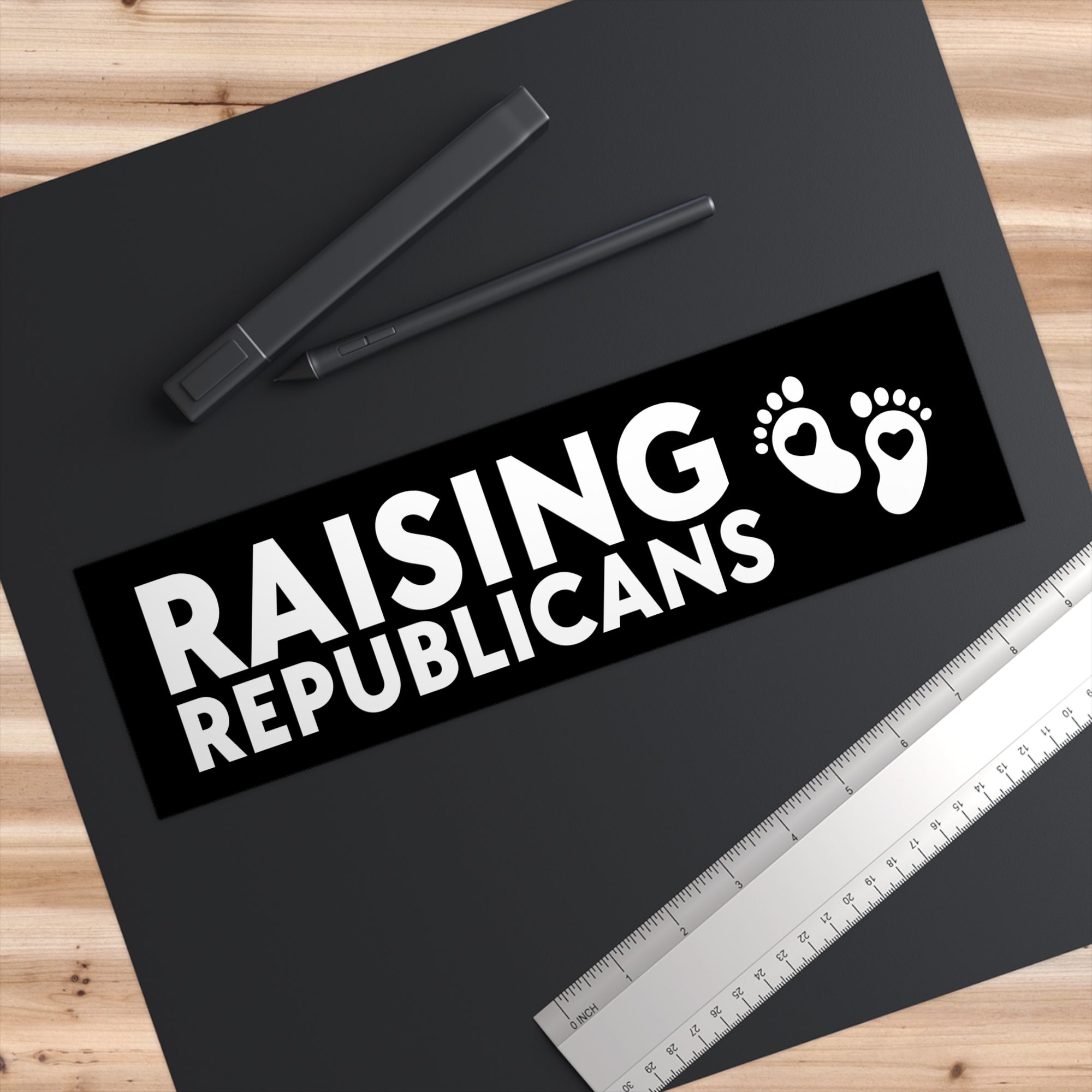 Sticker with 'RAISING REPUBLICANS' text and footprints on a black background, placed on a wooden surface with pens and a ruler.