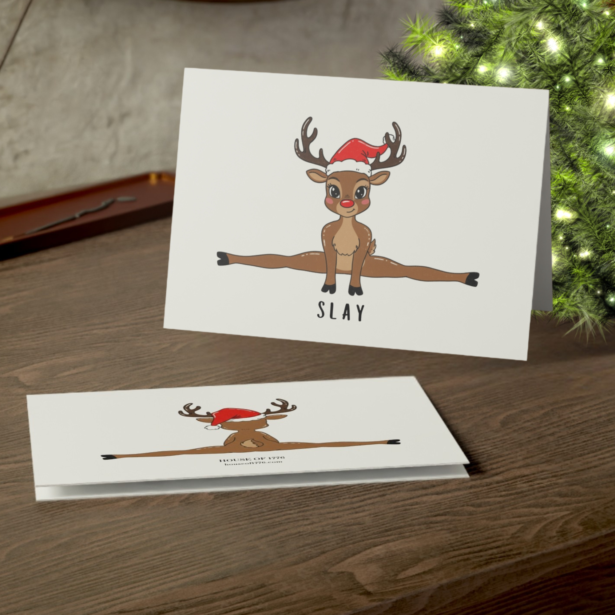 Card with a reindeer doing a split and the word 'SLAY' on a wooden surface with a Christmas tree in the background.