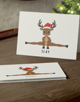Card with a reindeer doing a split and the word 'SLAY' on a wooden surface with a Christmas tree in the background.
