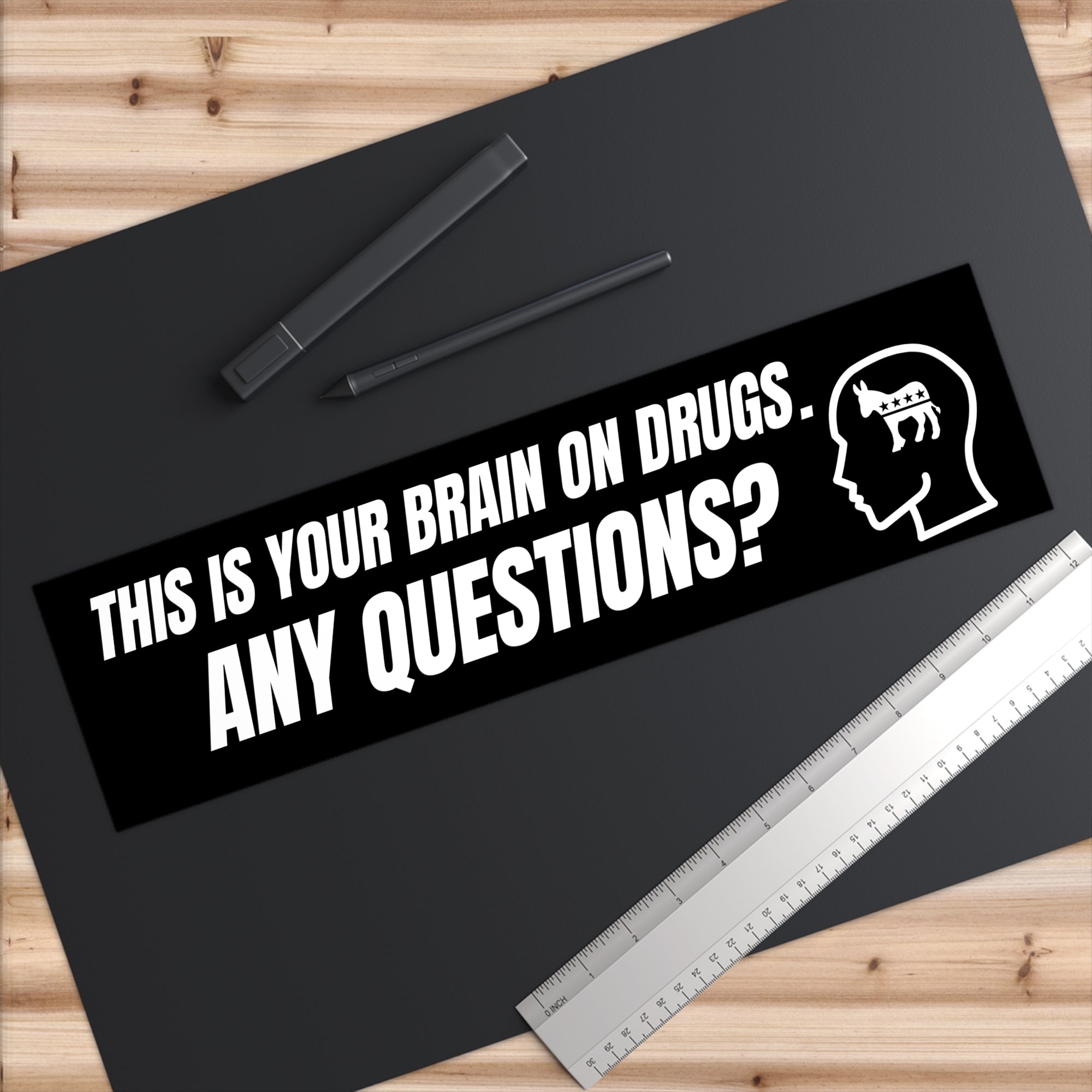 Sticker with text 'This is your brain on drugs. Any questions?' on a black surface with a ruler and pen.