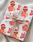 Vintage “Baby’s First Christmas” wrapping paper featuring a smiling dark-skinned baby girl in a pink romper and Santa hat holding a white bunny plush and candy cane, surrounded by red Christmas stockings and holly, retro storybook illustration style.