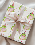 Gift box wrapped with a whimsical frog themed paper and a pink ribbon on a satin surface.