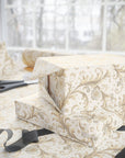 Gift box wrapped in wrapping paper featuring scroll work and flowers with a ribbon and scissors on a table.