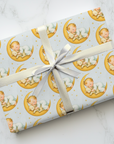 Gift wrapped in patterned paper with a baby on a moon design, tied with a ribbon on a marble surface.