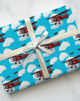 Wrapping paper with a pattern of red biplanes flying through a blue sky with white clouds.