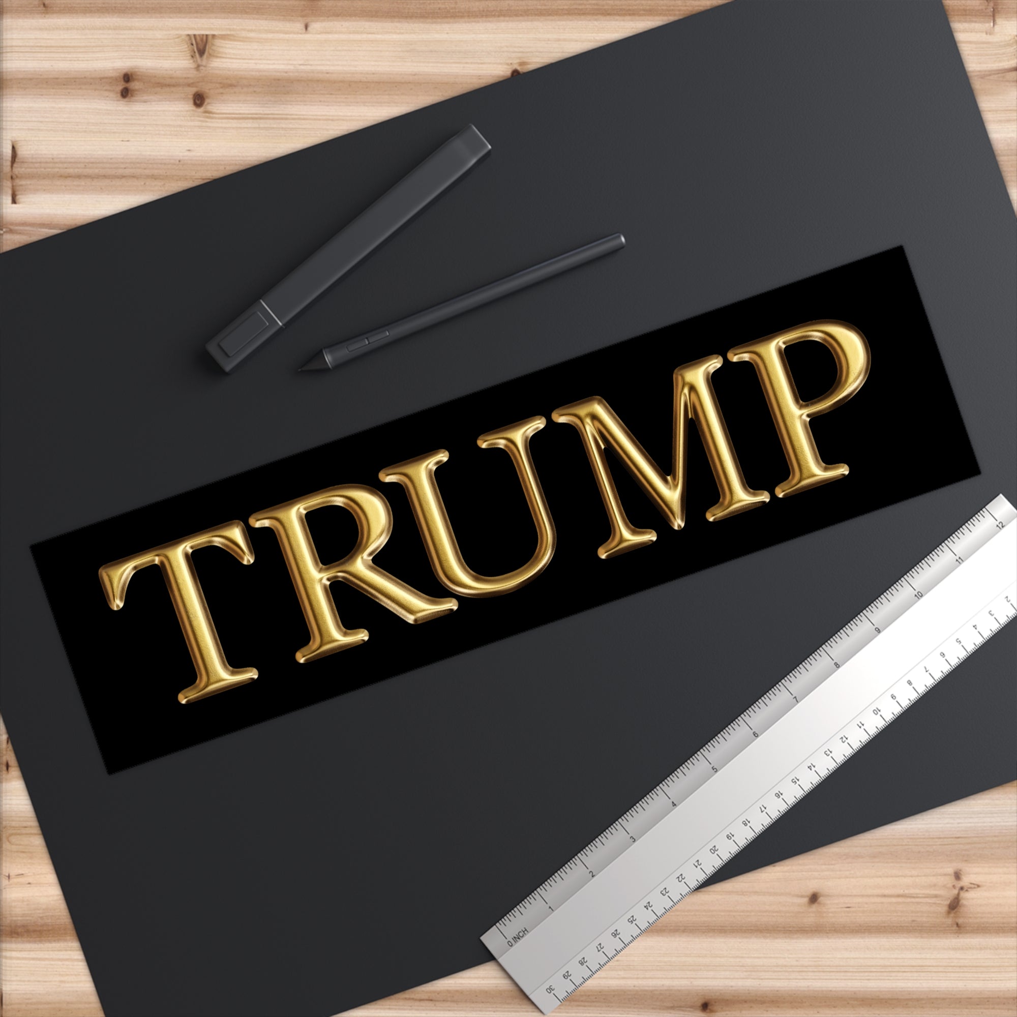 Gold 'TRUMP' text on a black background with a ruler and pens on a wooden surface