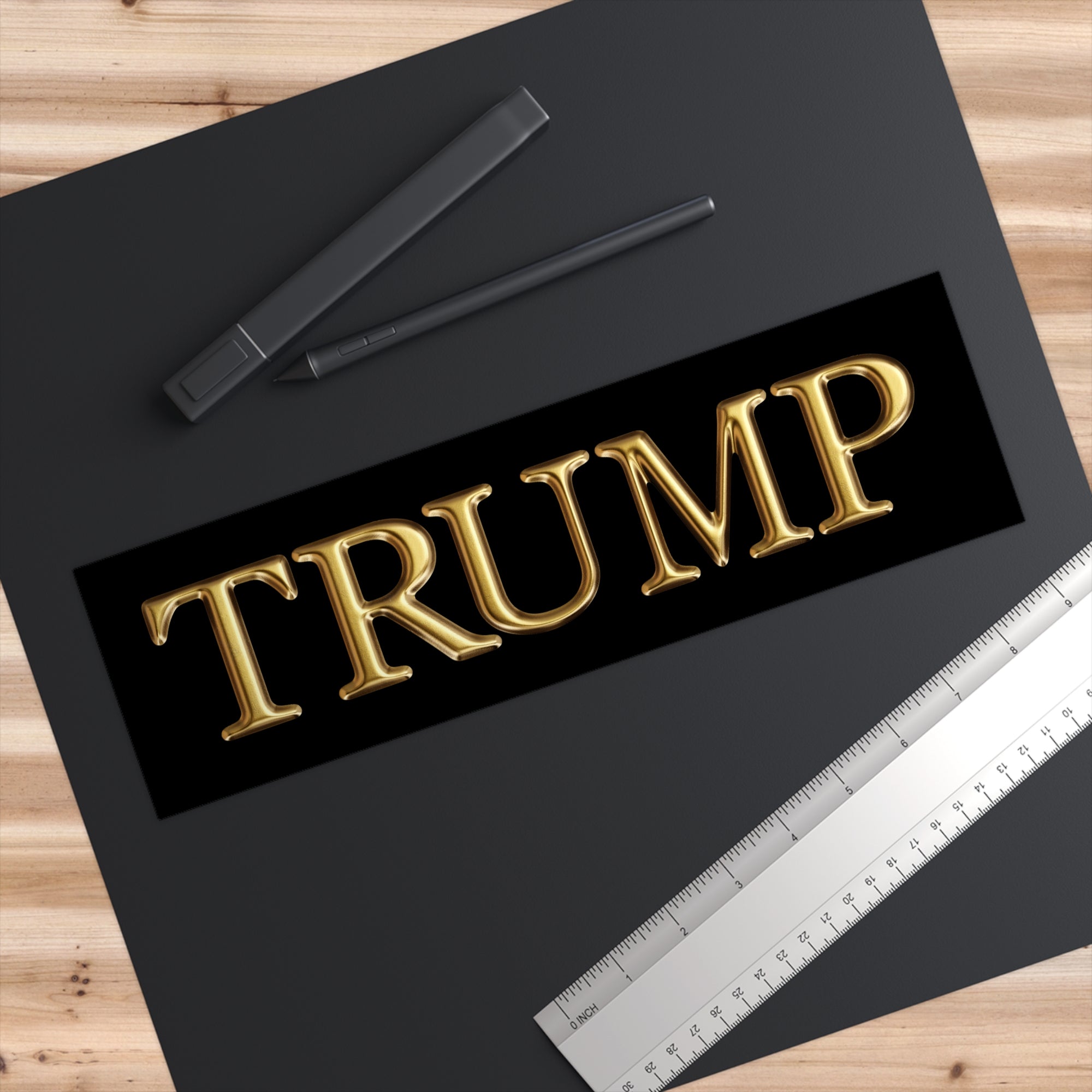 Gold 'TRUMP' text on a black background with pens and a ruler on a wooden surface
