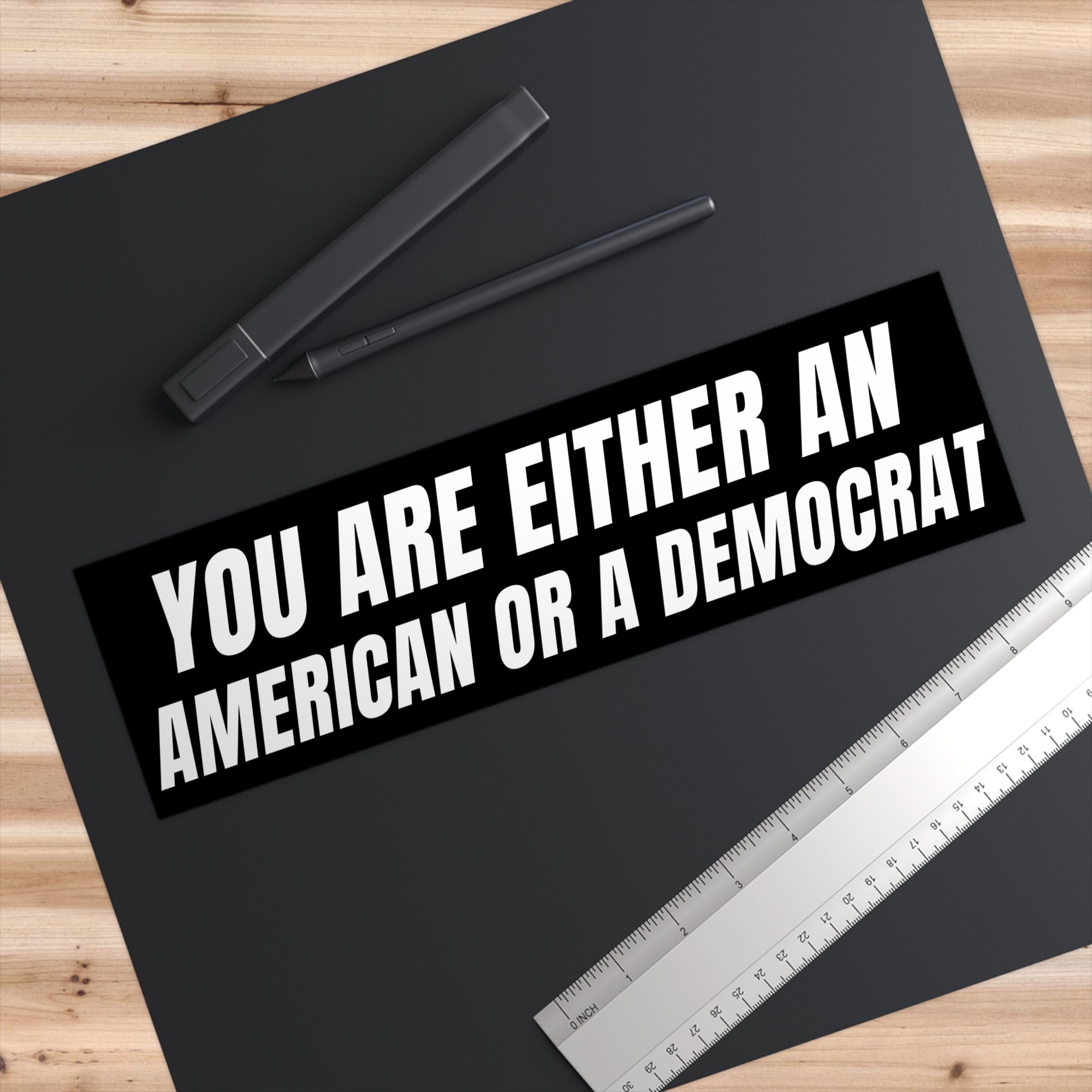 Black bumper sticker with text 'YOU ARE EITHER AN AMERICAN OR A DEMOCRAT' on a black surface with pens and a ruler.